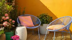 Image of an outdoor patio with bright yellow walls and lots of plants in the corner. There are two powder blue accent chairs with yellow pillows on them.