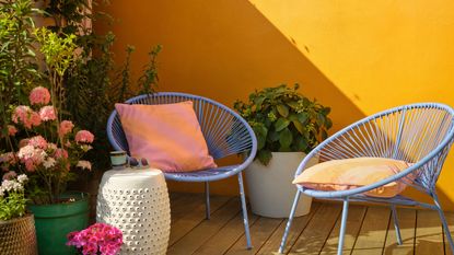 Image of an outdoor patio with bright yellow walls and lots of plants in the corner. There are two powder blue accent chairs with yellow pillows on them.