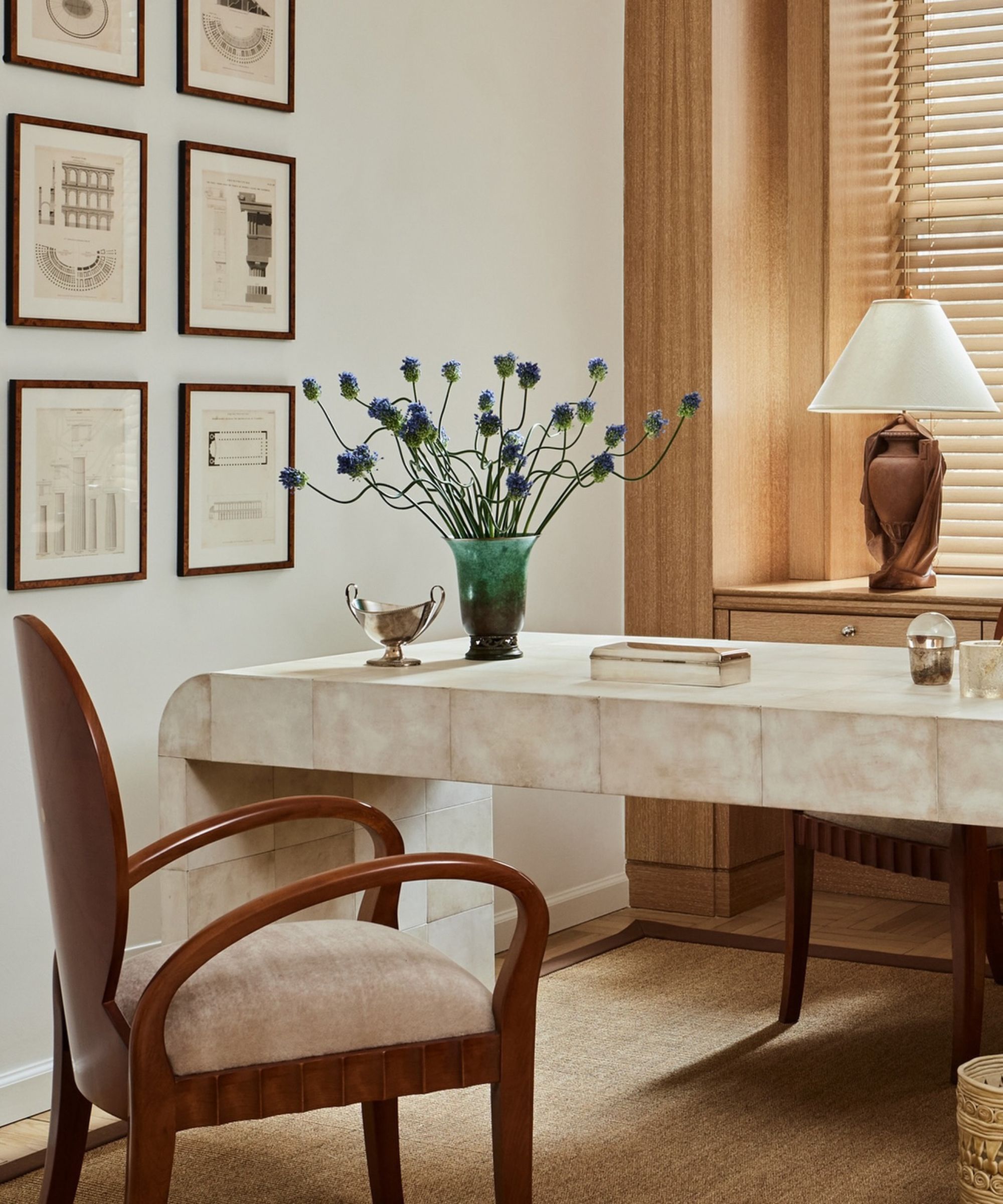 Large curvacous stone desk, vase of flowers on table, wooden chair placed in-front, artwork on walls, lamp behind