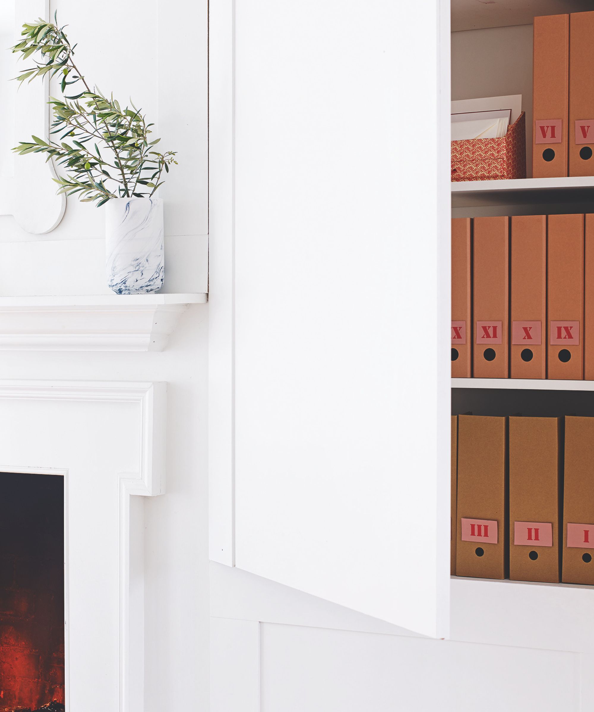 A white storage cupboard filled with paper files in folders, next to a white fireplace with a white and blue base on top with green branches in it.