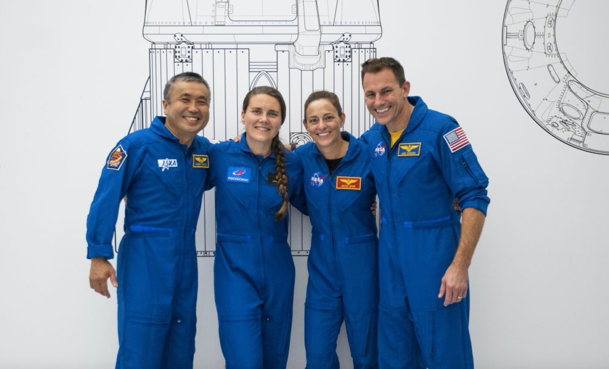 four blue suited astronauts in front of a wall with space design drawings