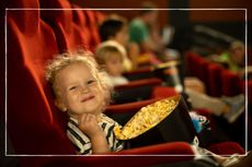 Curly haired child smiling at the camera as she sits in a cinema seat, with a large bucket of popcorn in her lap