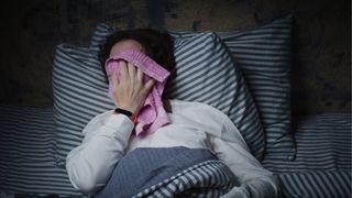 A woman in bed with navy and white stripey sheets holding a pink flannel over her face to cool down, wearing white pyjamas and a red smart watch