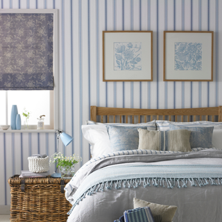 Blue and white bedroom with striped wallpaper, floral roller blind, wicker basket bedside table, knitted white baskets, bedlinen