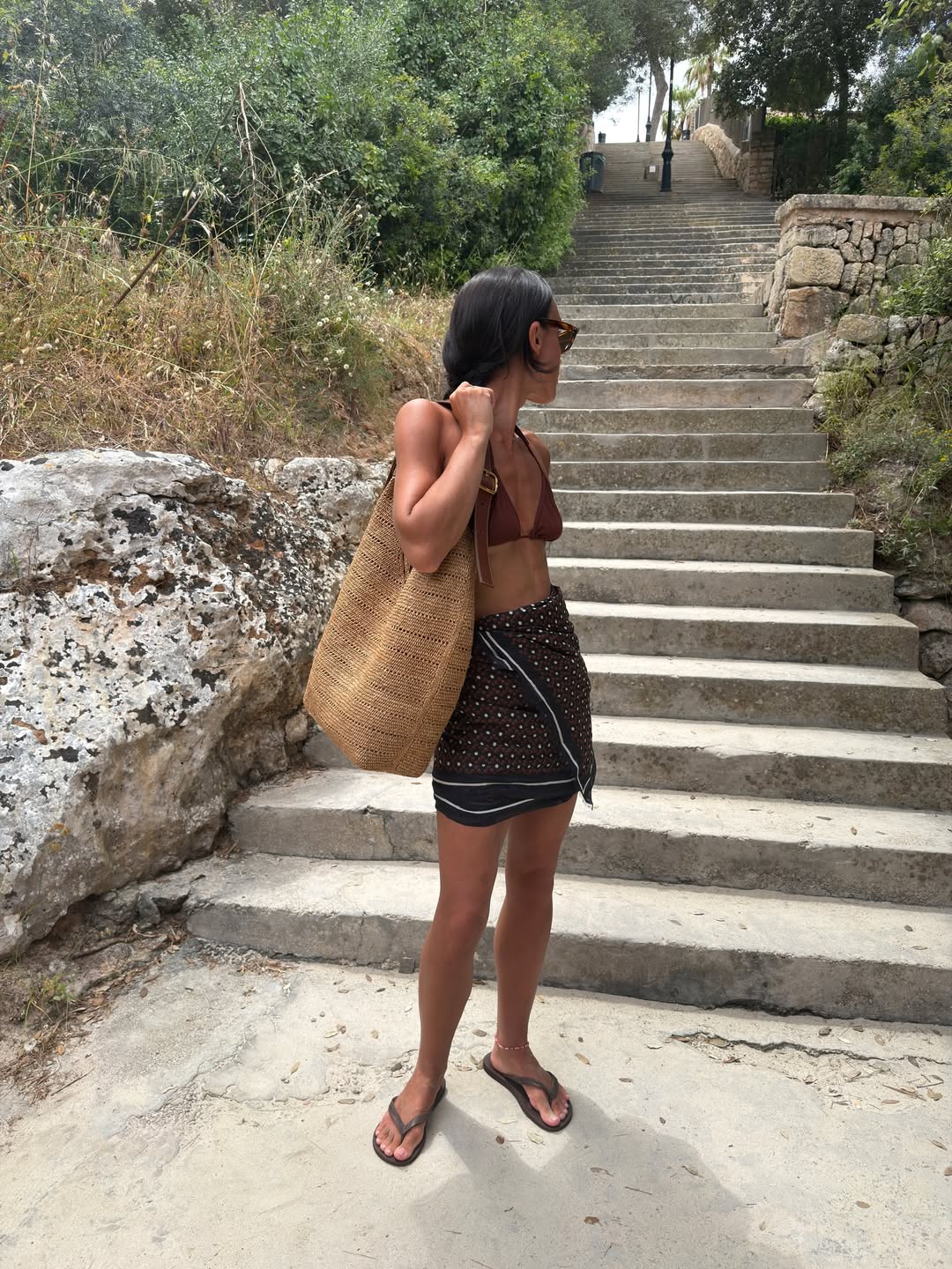 EMily Wells wears a brown bikini top, mini wrap skirt, sandals and a beach bag