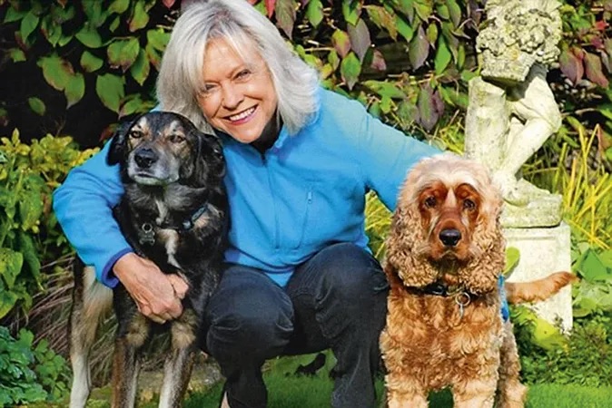 Sue Barker kneeling in a garden between two dogs &amp;mdash; a black mixed-breed dog and a golden cocker spaniel &amp;mdash; smiling at the camera beside greenery and a stone garden ornament.