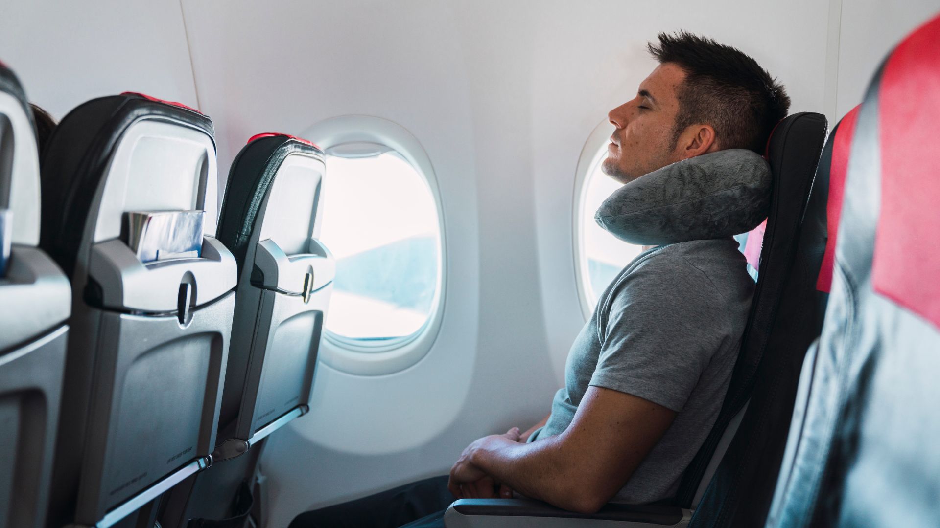A man sleeping on plane with a grey neck pillow