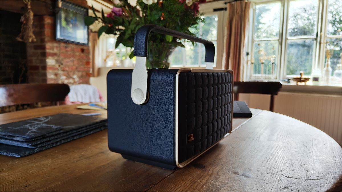 A side angle of a black JBL Authentics 300 wireless speaker. It is positioned on a wooden table with a plant in the background. The handle on the speaker is extended upwards.