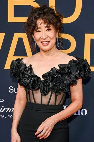 Image of Sandra Oh smiling, with a curly brunette bun and wearing a black floral dress