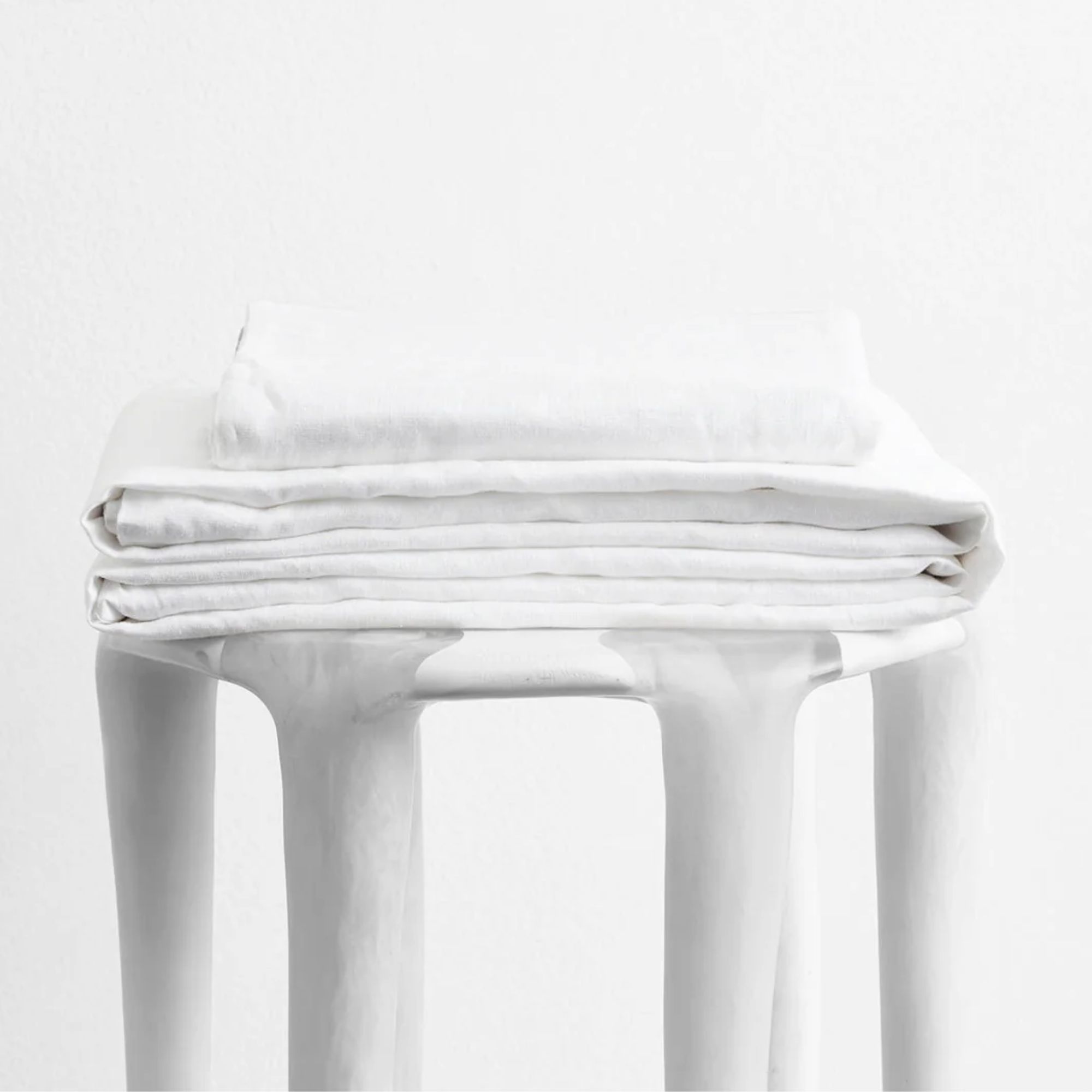 A folded BedThreads White Sheet Set on a white stool against a white background.