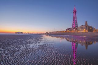 A view of Blackpool seafront, taken from the beach. The sand is wet, and there is a golden sunrise on the horizon. Blackpool Tower is lit up with magenta lights.