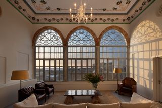 A lounge space with Ottoman Empire features. Three arched windows cover the entire far wall. The space is filled with a large table with a vase of flowers on it, in the centre, surrounded by leather armchairs in deep brown and a beige sofa.