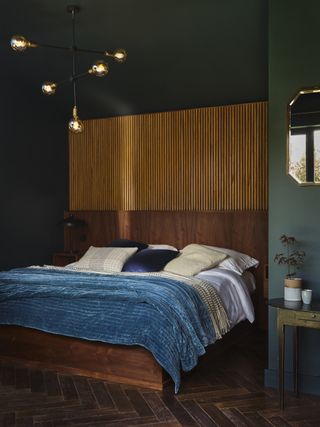 green bedroom with wooden slats behind wooden bed, blue blanket, pendant light, mirror