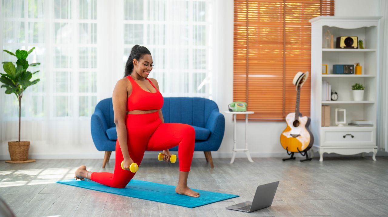 A woman in a sports bra and leggings works out in a living room. She is in a low lunge on a yoga mat with a pair of dumbbells in her hands. Her left knee is in front of her body, bent at a right angle, and her left foot placed on the floor. Her right knee and shin are behind her, with her right foot pointing backwards. Behind her we see a guitar, decorative shelving, a sofa and a leafy plant.