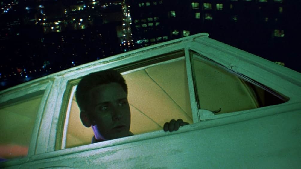 Screenshot from the movie Repo Man. A startled-looking man is poking his head out of a car window. The car is glowing in the dark and flying high above the city.