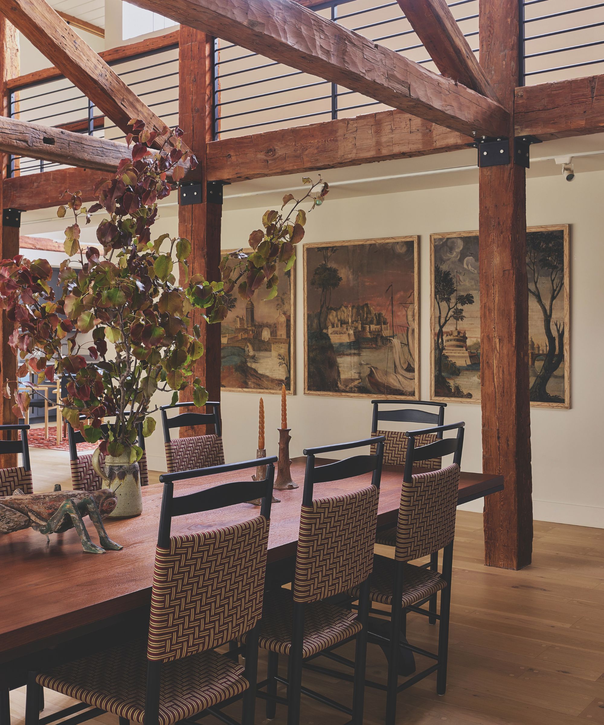 Rustic wooden dining table surrounded by woven-back chairs under heavy timber posts, featuring framed artwork and leafy branch decor.