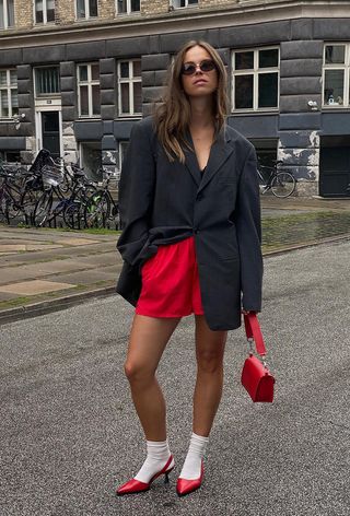 A woman wearing a gray oversize blazer with red basketball shorts, white socks, red slingback heels, and a red shoulder bag.