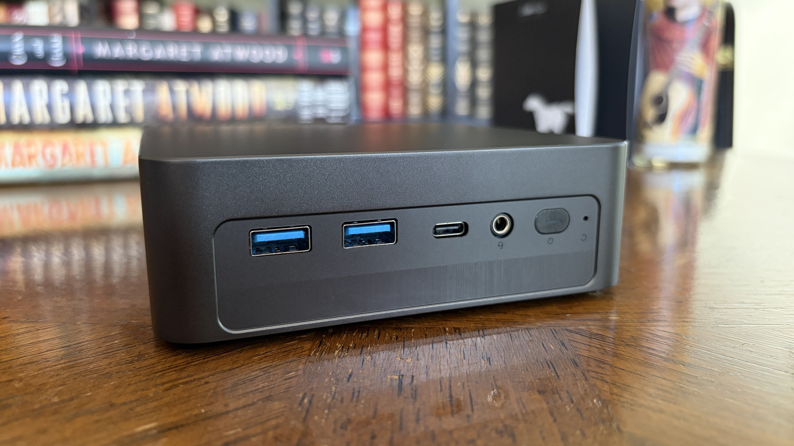 Close-up of a compact Chuwi mini PC with USB, USB-C ports, audio jack, and power button, set against a bookshelf background.