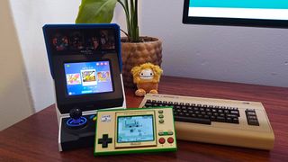 Neo Geo Mini on left, C64 mini on right, and Zelda: Game and Watch in centre on woodgrain desk with plant in backdrop next to Big Foot Squishmallow.