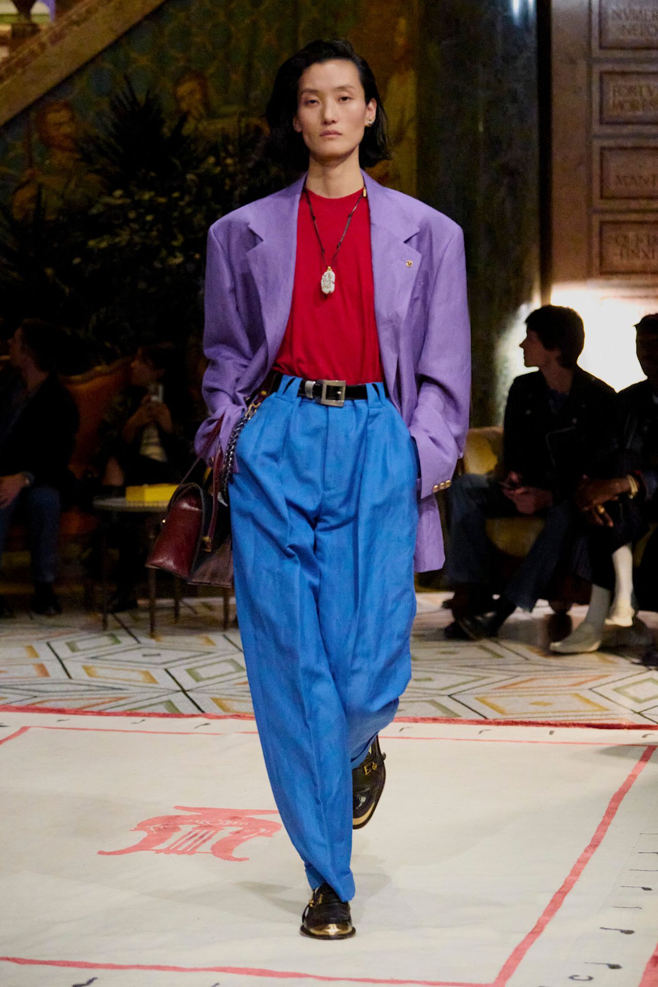 A Versace model wearing a purple blazer layered on top of a red crewneck shirt, bright blue trousers, black oxfords, a cord necklace, a red handbag, and a black belt.
