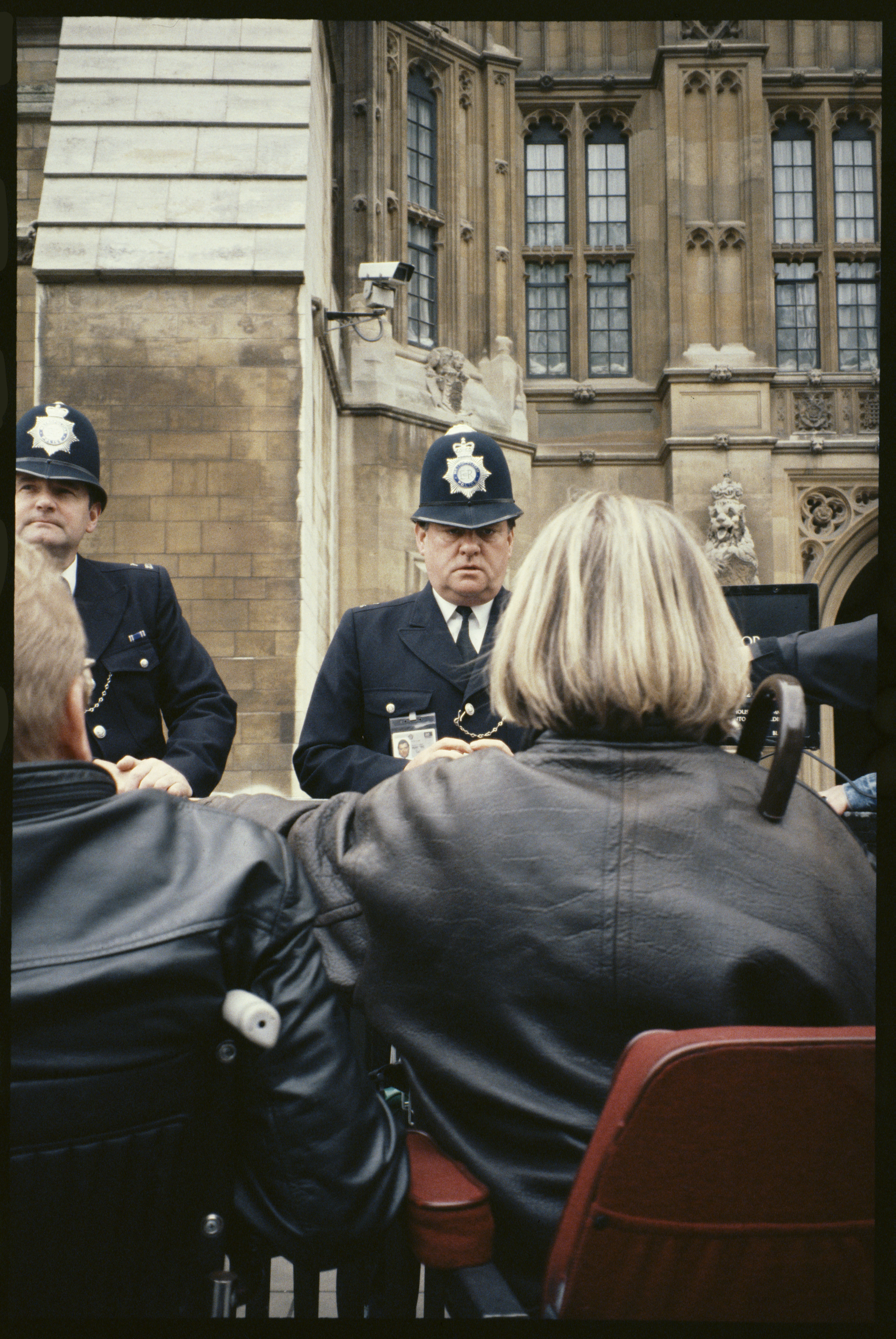 Two Metropolitan Police officers face a group of wheelchair users outside the Houses of Parliament, the Gothic stonework and a CCTV camera visible above them.
