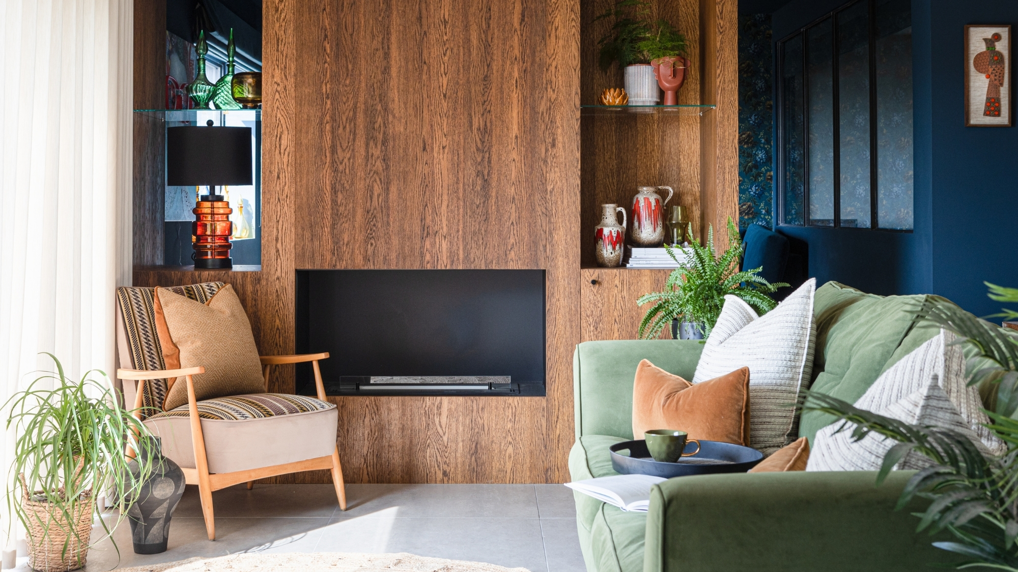 Green velvet sofa, patterned armchair, wood-panelled wall with fireplace, colourful pottery and plants on shelves.
