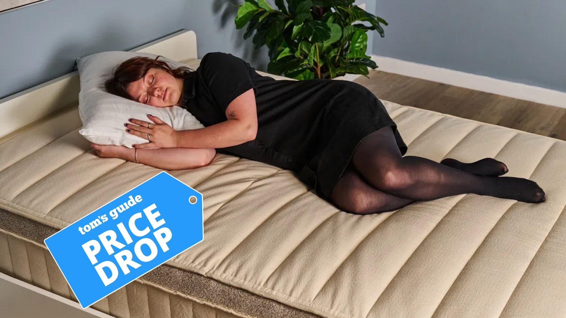 The image shows a dark haired woman wearing a black dress and black tights lying on her side on a beige mattress, placed in a blue bedroom. Tucked under her head is a white pillow.