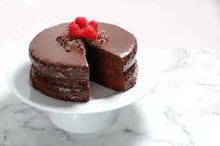 A chocolate cake with raspberries on top on a cake stand in a kitchen.