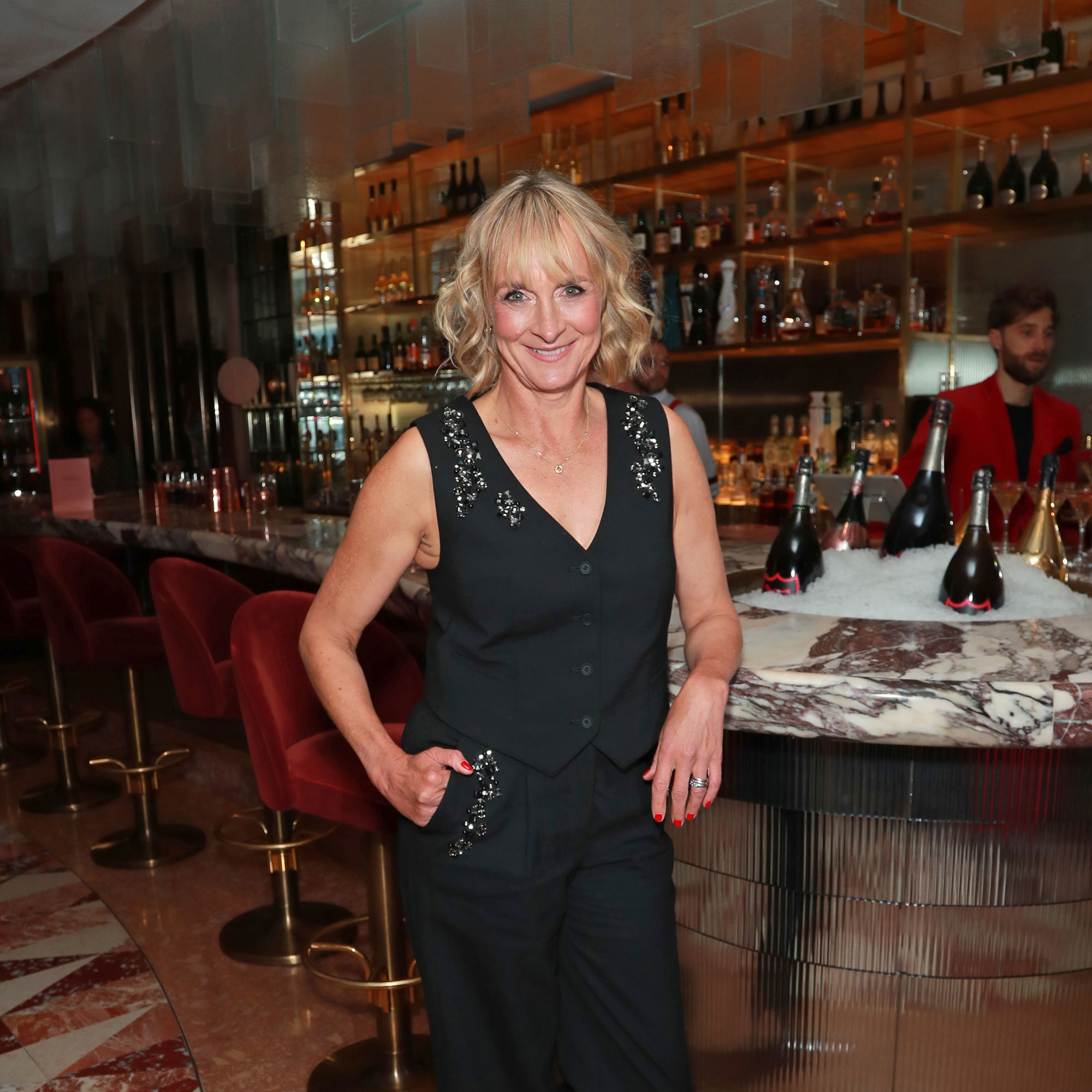 Louise Minchin in a black waistcoat and trousers leaning on a marble bar