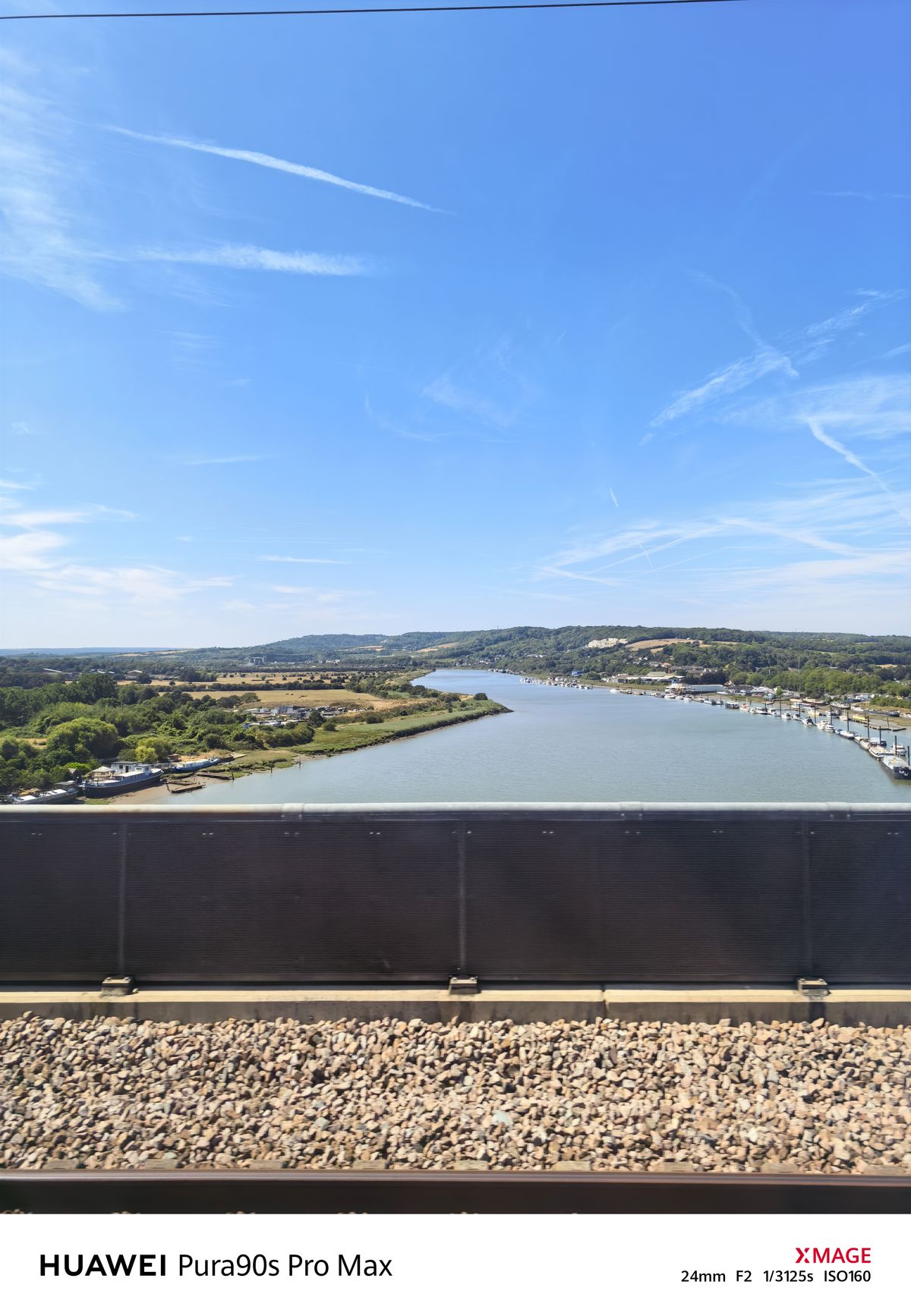 View across a wide river toward green hills and a marina, with railway tracks and a barrier in the foreground.