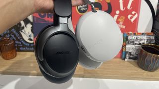 A hand holding the white Sonos Ace and black Bose QuietComfort Ultra Headphones by their headbands in front of some vinyl sleeves.
