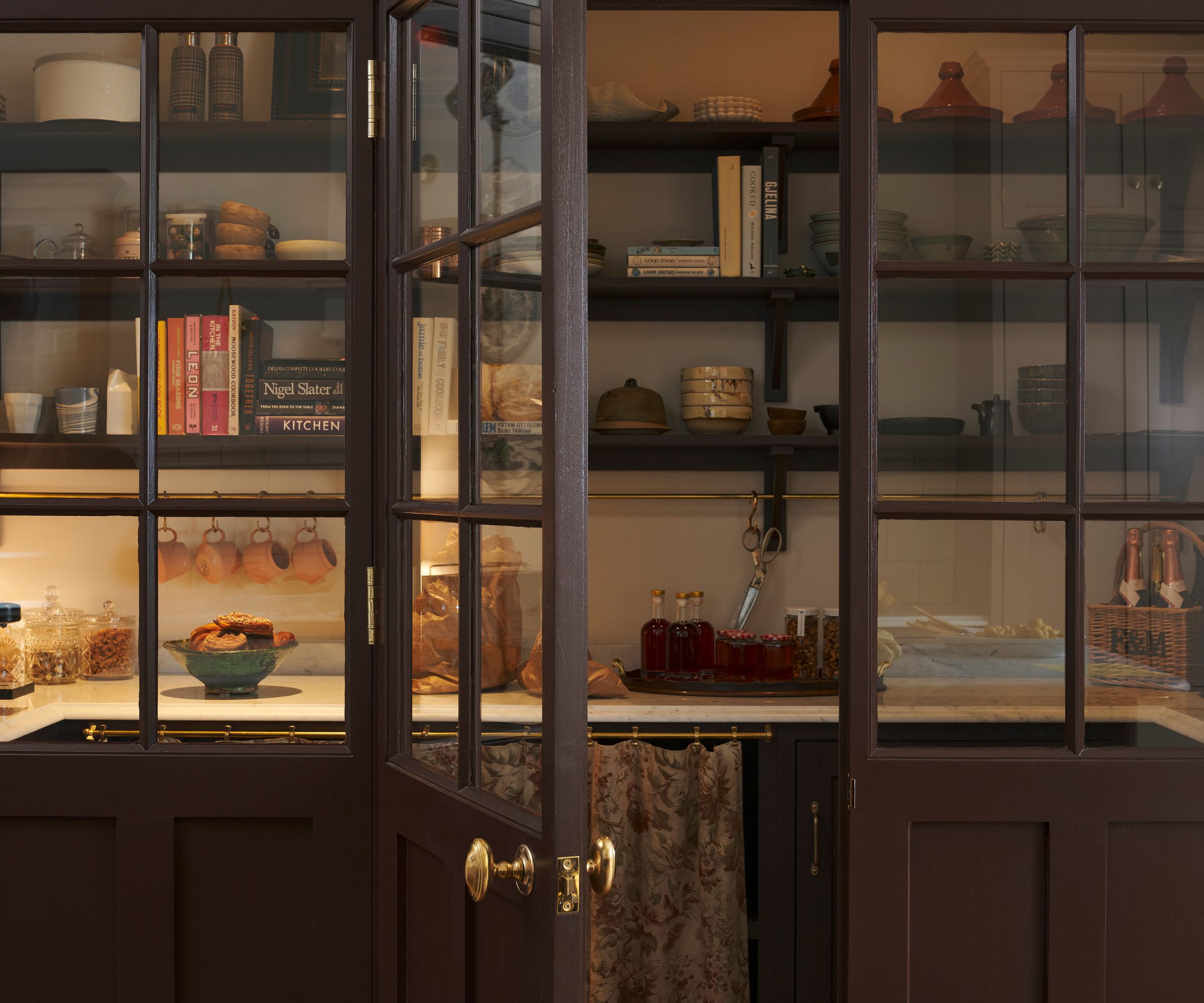 A walk-in pantry with brown and glass doors, open shelving on the walls, cabinets below, and a cabinet skirt over part of the cabinetry