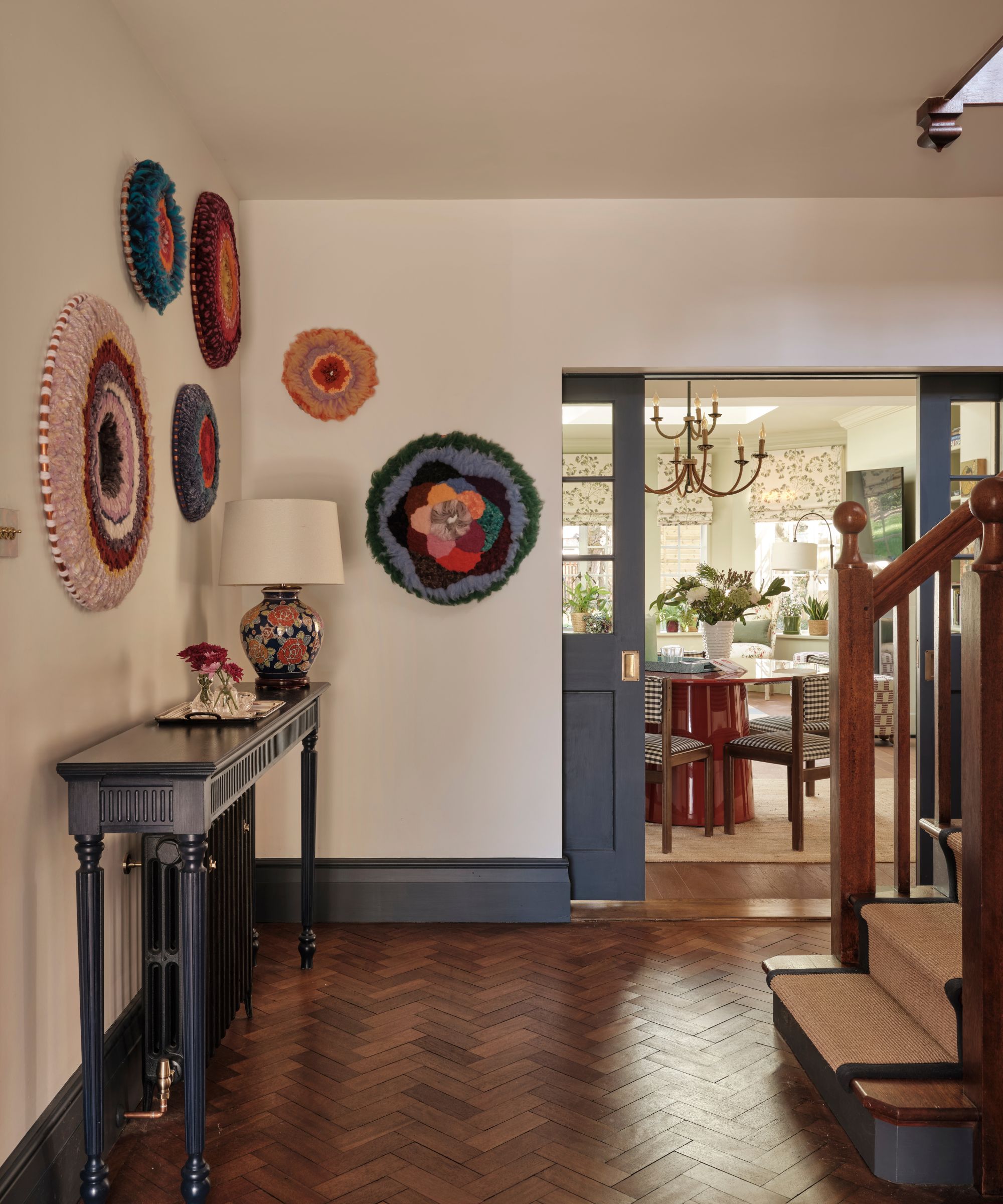 Entrance hallway with herringbone wood flooring, a dark wood console table, and colorful circular woven wall hangings; a glimpse into a floral-themed living area beyond