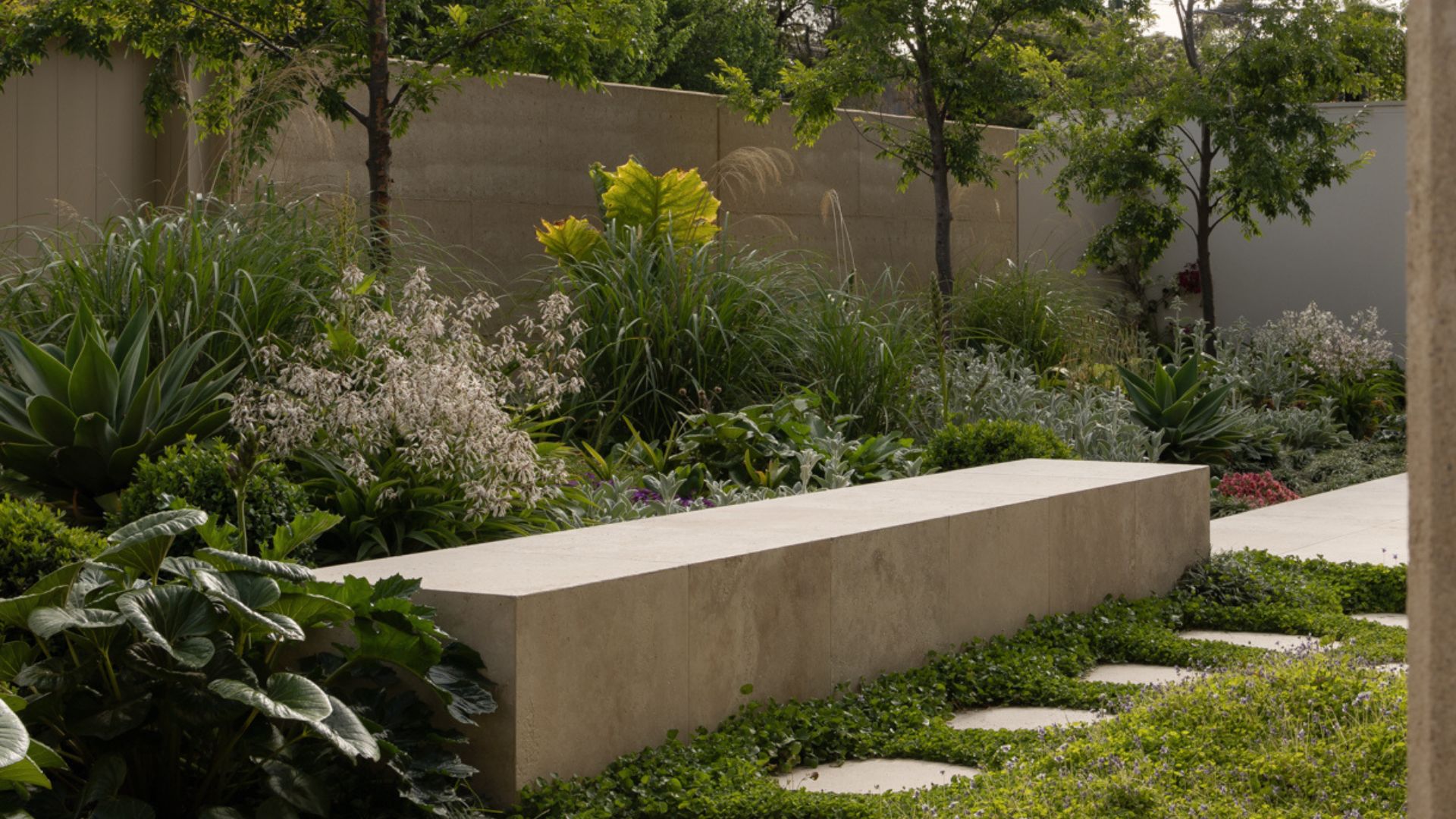 An outdoor garden with a stone bench, a path with ground cover and textured plants and trees around the zone