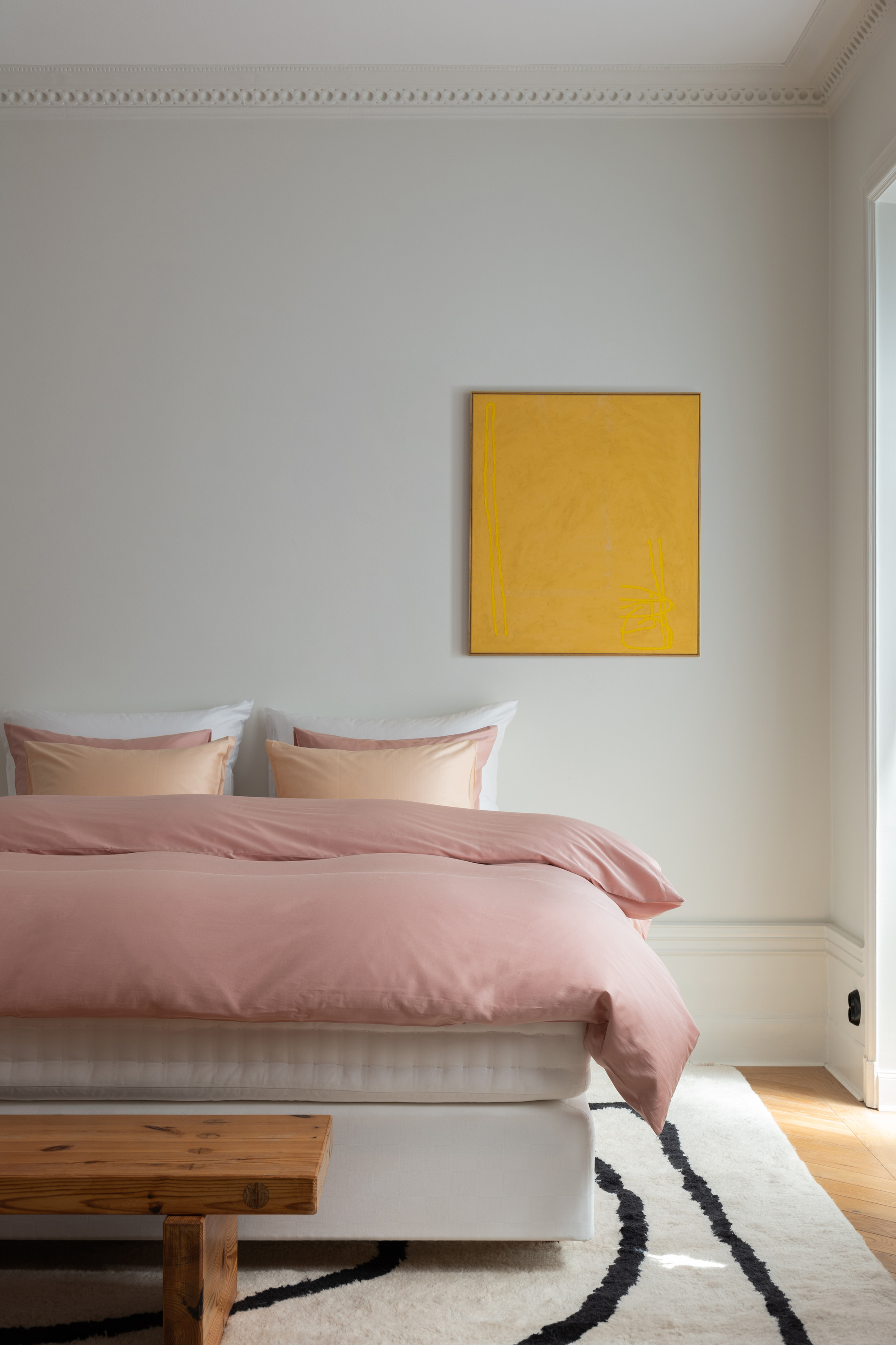 A bedroom with white walls, pink bedding, and a yellow canvas on the wall
