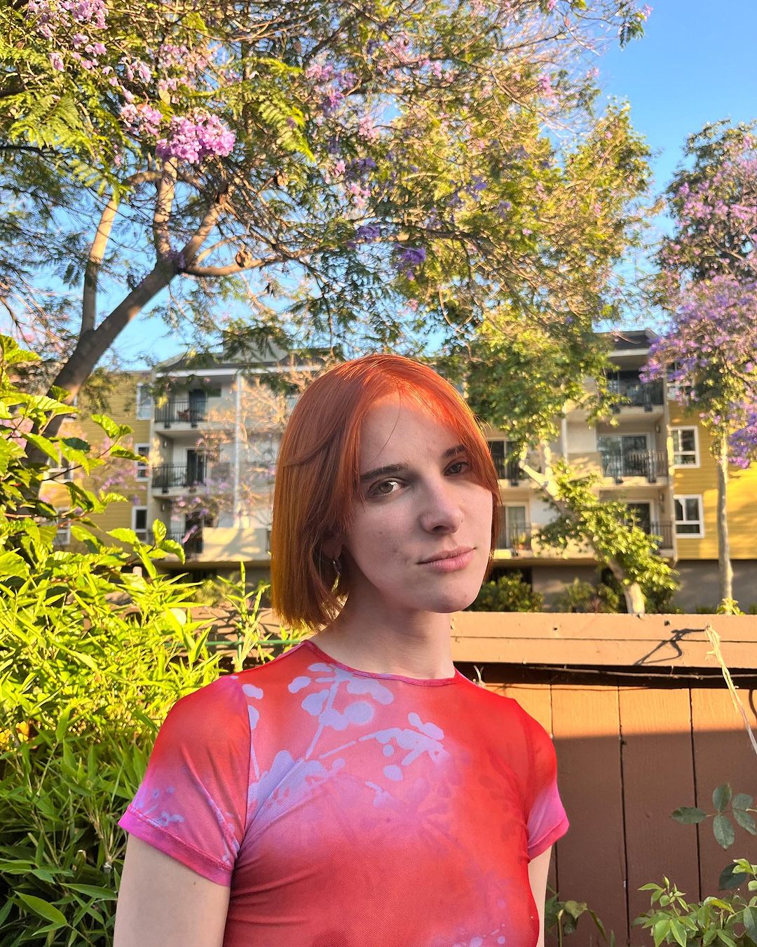 A woman, Hari Nef, with short red hair gazes at the camera surrounded by trees.