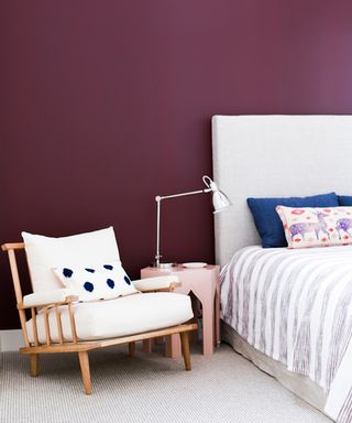 Aubergine wall, relaxed lounge chair with indigo spot cushion, pink bedside with chrome lamp, and striped bedding with colored pillows.