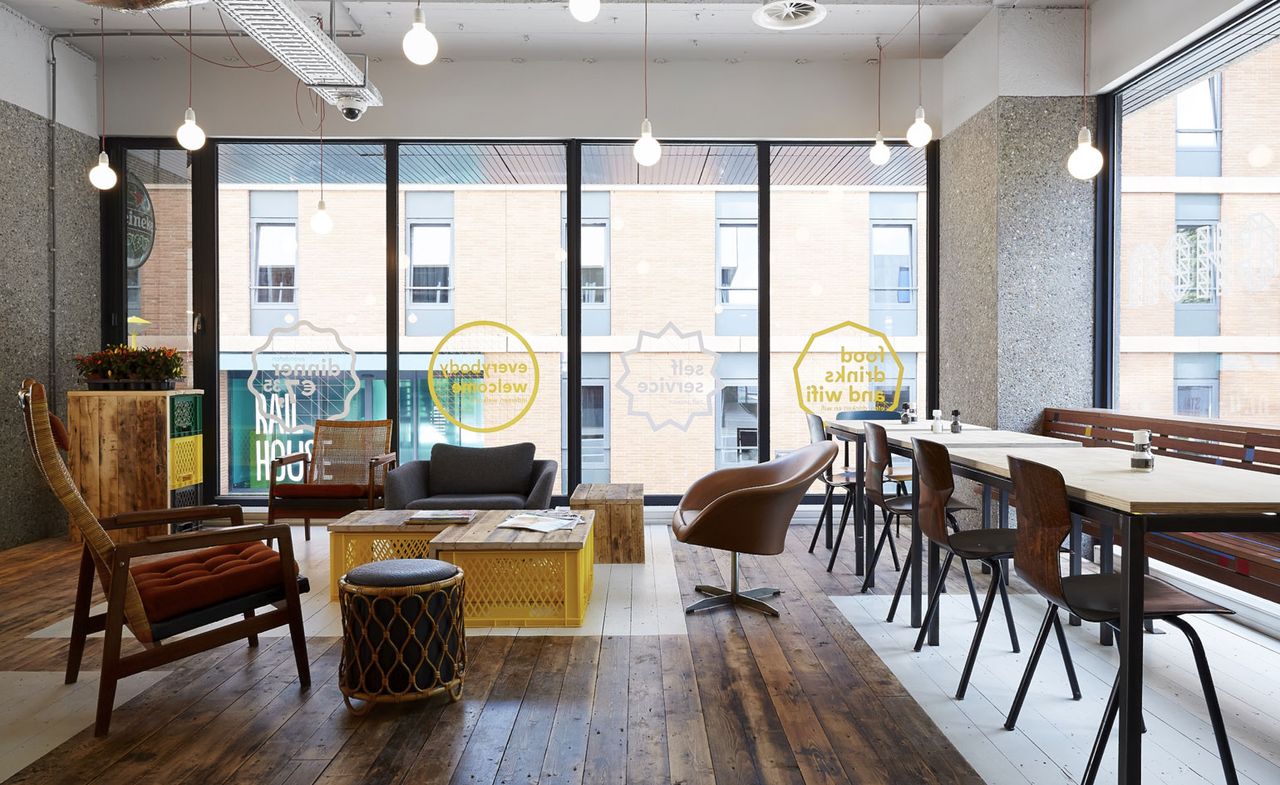 Restaurant in the Meininger Hotel. Open and bright space, with floor-to-ceiling windows. Wooden benches and chairs serve as seating, with coffee tables that are made out of plastic boxes topped with wooden boards.