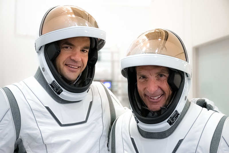 Two men in white space suits are shown from the chest up, with their copper visors raised to reveal their faces hidden inside their helmets, laughing in a white room.