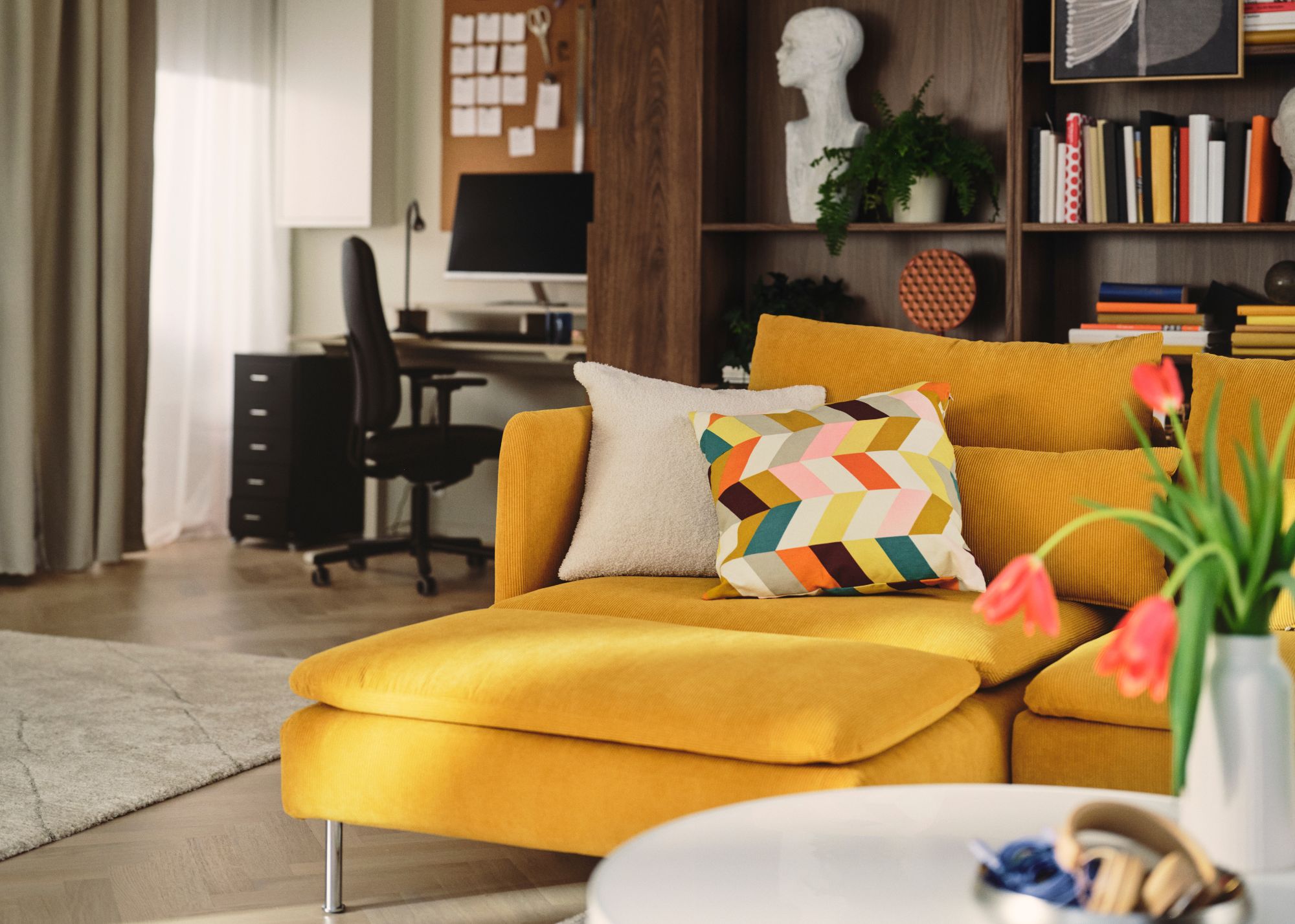 A living room with a yellow sofa with chaise longue in front of dark wood shelving with books and decor objects, with a desk area in the back corner of the room