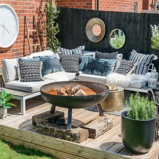 garden with a decking area with a grey corner sofa, fire pit, potted plants, circular white clock and grey fence