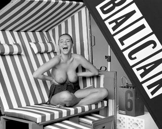 Black & white photograph of a laughing woman with her check bare, sitting on a piece of beach furniture.