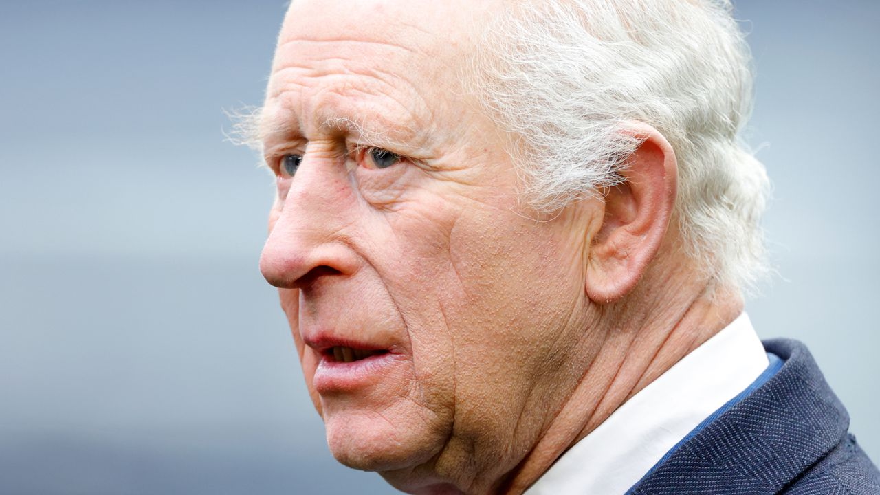 A headshot of King Charles wearing a blue suit and white shirt turned to the left and looking serious