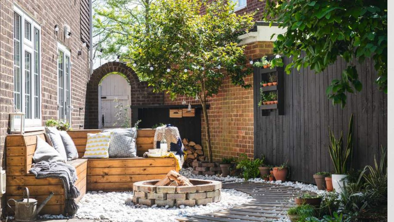 Small garden with tree, wooden outdoor seating, fire-pit and wooden boardwalk on white gravel, black wooden fence.