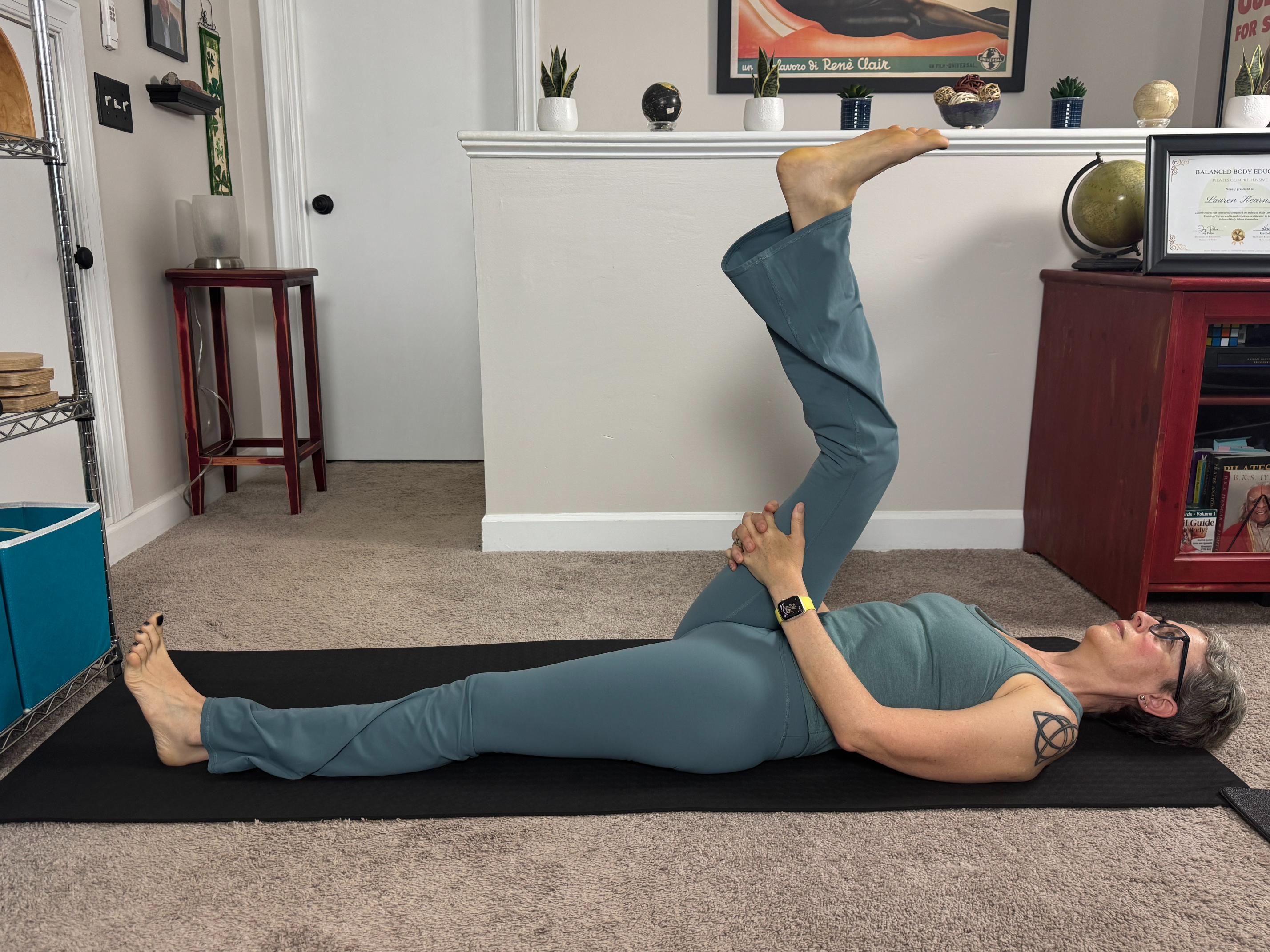 woman lying on her back on a black yoga mat in a living room performing a leg stretch with one leg extended and the other bent