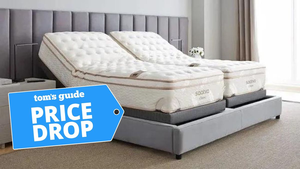 Image shows a split-king Saatva Classic mattress on a grey quilted bed frame in a light grey bedroom, with a blue Price Drop sales badge overlaid