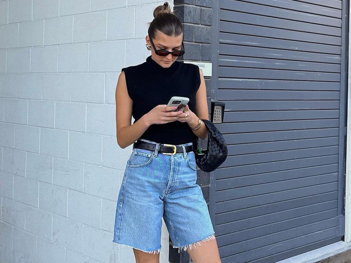 female fashion influencer Sara Walker looks at her Iphone cell phone in front of a white cinder brick wall wearing a slicked back bun, black oval sunglasses, a mock neck black sleeveless top, a mini black Bottega Veneta bag black belt, and Bermuda denim cutoff shorts