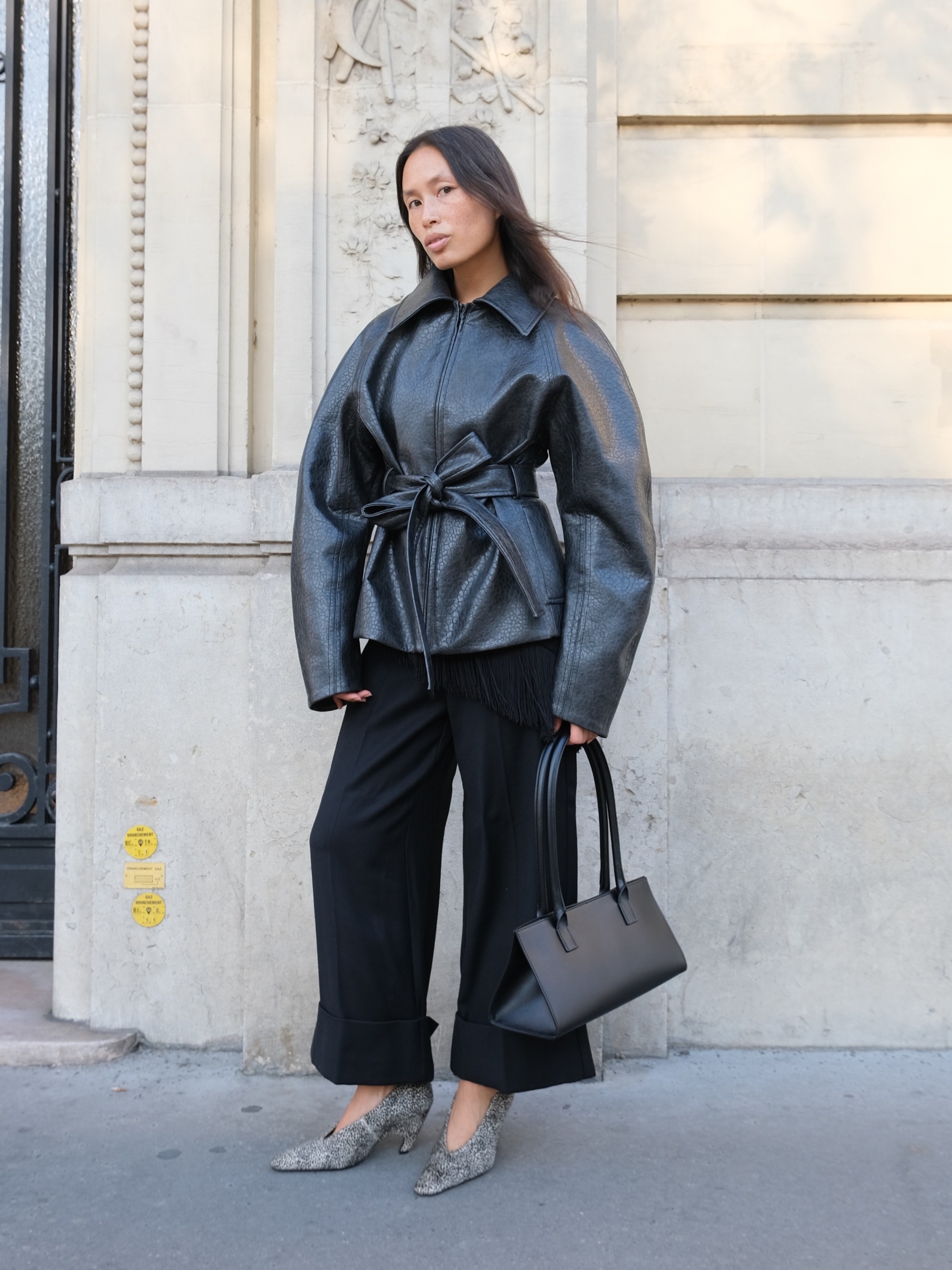 An image of an influencer in Paris wearing a leather waisted jacket with black wide-leg trousers and snake-print heels holding a black handbag