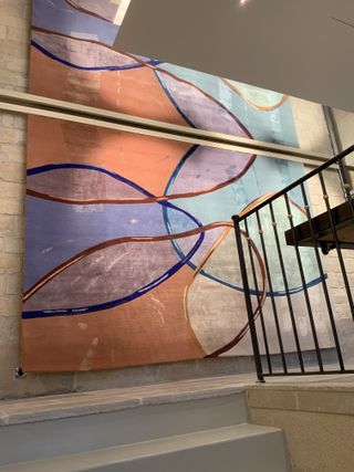 Interior view of Deirdre Dyson’s showroom featuring a large peach, purple and blue rug hanging on a stone wall by the stairs