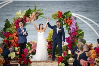 Ellie and Ben cheer as they get married on the beach with a flower arch behind them.